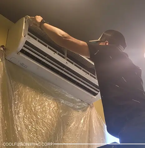Technician cleaning an air conditioning unit mounted on a wall, with protective plastic covering the area below